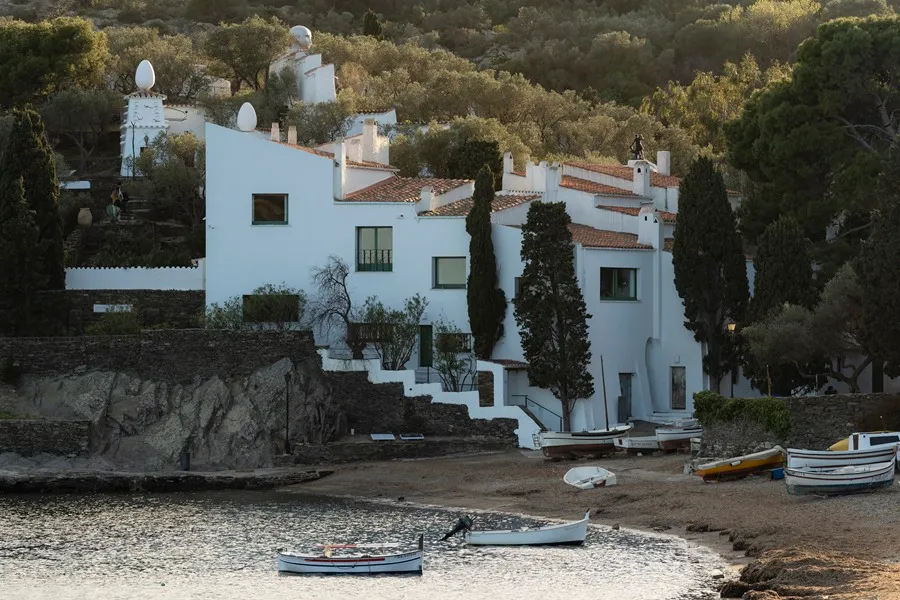 Vista exterior de la fachada blanca de la Casa-Museo Salvador Dalí en la bahía de Portlligat, Cadaqués, con pequeñas barcas de pesca tradicionales varadas en la arena y las esculturas de huevos surrealistas en los tejados rodeadas de olivos.