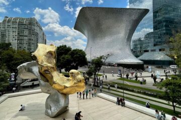 Museos gratuitos de CDMX: una guía imperdible Vista panorámica exterior del Museo Soumaya en Plaza Carso, Ciudad de México, tomada desde la terraza del Museo Jumex, mostrando la arquitectura plateada de Fernando Romero y esculturas modernas al aire libre bajo un cielo parcialmente nublado.
