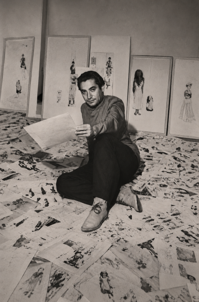 Fotografía histórica en blanco y negro de Ramón Valdiosera joven, sentado en el suelo de su estudio de arte rodeado de ilustraciones y bocetos de moda indígena mexicana.