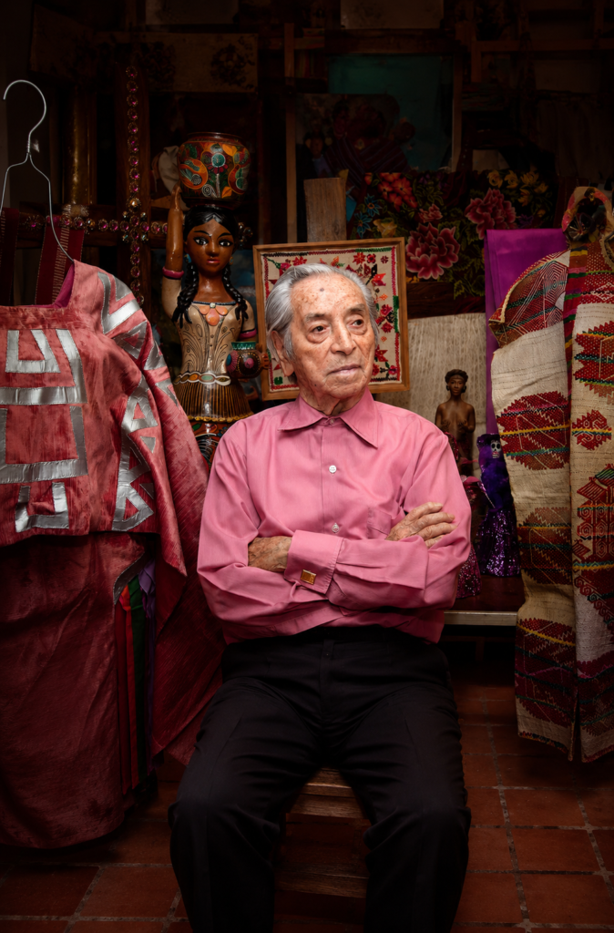 Retrato vertical de estudio de Ramón Valdiosera sentado con los brazos cruzados, vistiendo una camisa rosa intenso frente a textiles tradicionales de su acervo de moda mexicana.