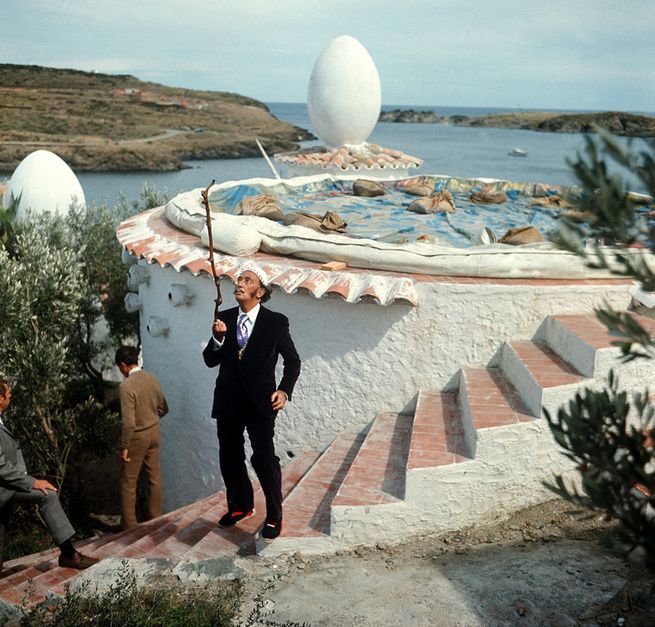 Salvador Dalí con traje sosteniendo un bastón en las escaleras exteriores de su casa de Portlligat en Cadaqués, con esculturas de huevos gigantes surrealistas en el tejado y el mar de la Costa Brava al fondo.
