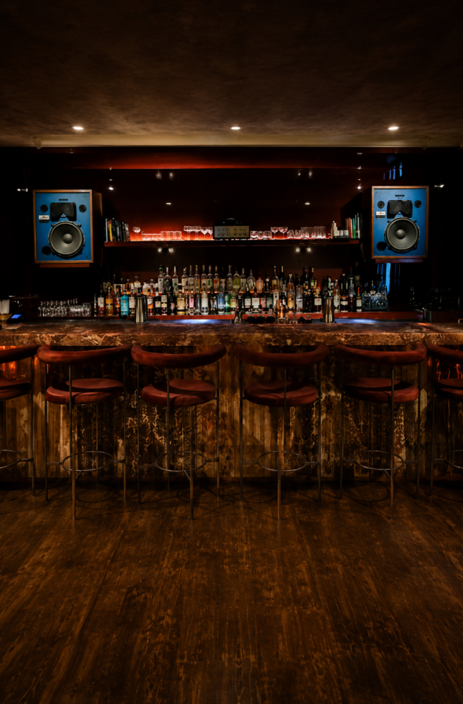 Barra de bar con asientos de terciopelo marrón en Shhh Listening Bar de la Ciudad de México, con estanterías de licores iluminadas y grandes altavoces antiguos JBL de color azul a los lados.