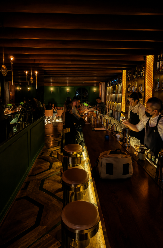 Barra de bar elegante en Tokyo Music Bar CDMX con taburetes dorados, paredes verde esmeralda, lámparas de banquero y un gran estante iluminado lleno de discos de vinilo.