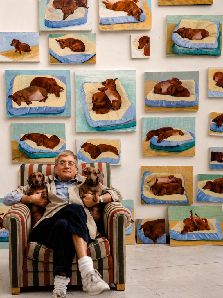 El artista británico David Hockney sentado en un sillón a rayas en su estudio de Los Ángeles, sosteniendo a sus dos perros salchicha (teckels) de color marrón, Stanley y Boodgie, rodeado de una pared repleta de sus pinturas al óleo de la serie Dog Days.