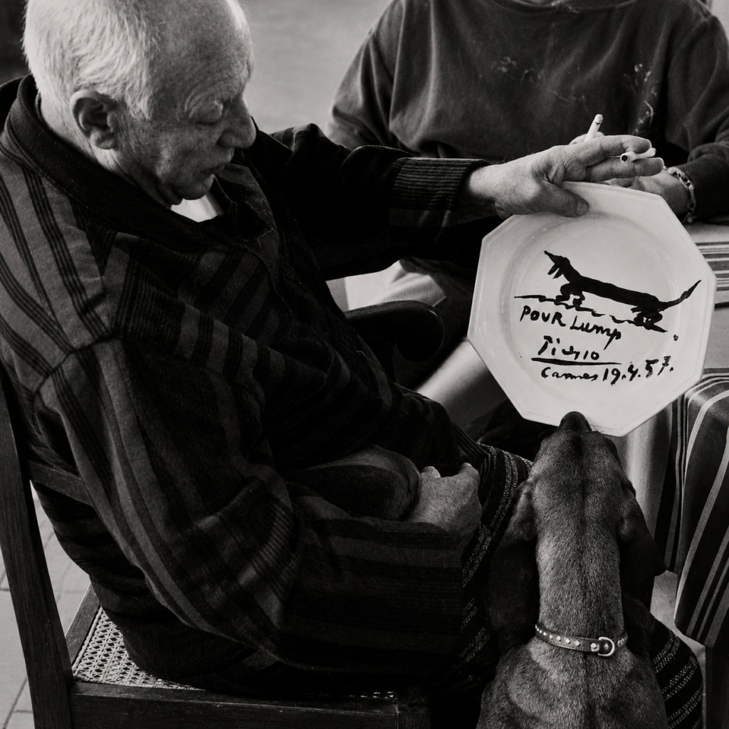 El artista español Pablo Picasso en su residencia de Cannes mirando un plato de cerámica blanca recién pintado con la silueta de su perro salchicha Lump, en una fotografía en blanco y negro tomada por David Douglas Duncan.