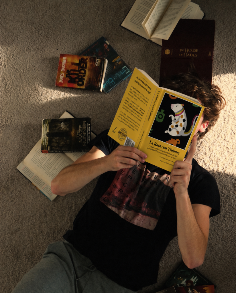 Persona acostada en una alfombra leyendo el libro La Rata con Thinner de Omar Ramírez rodeada de novelas juveniles como The Maze Runner y The House of Hades.