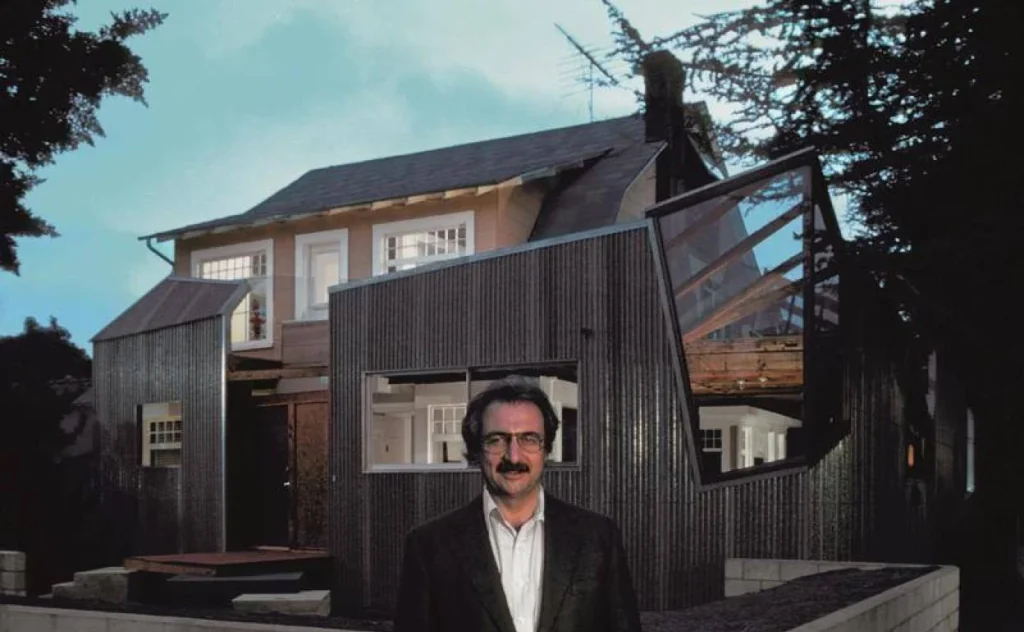 Retrato histórico del arquitecto canadiense-estadounidense Frank Gehry posando en 1978 frente a la fachada de su revolucionaria e icónica residencia deconstructivista de metal corrugado y madera en Santa Mónica, California.