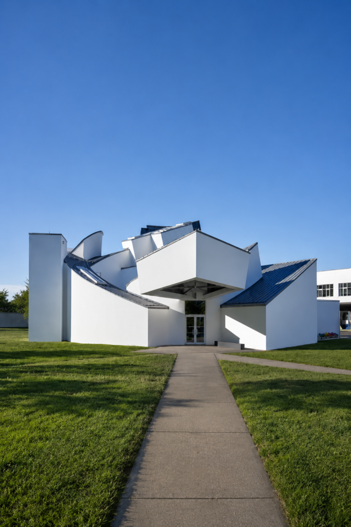 Vista exterior diurna y simétrica del sendero que conduce al Vitra Design Museum en Weil am Rhein, Alemania, obra maestra de la arquitectura deconstructivista de yeso blanco y cubiertas de zinc diseñada por Frank Gehry en 1989.