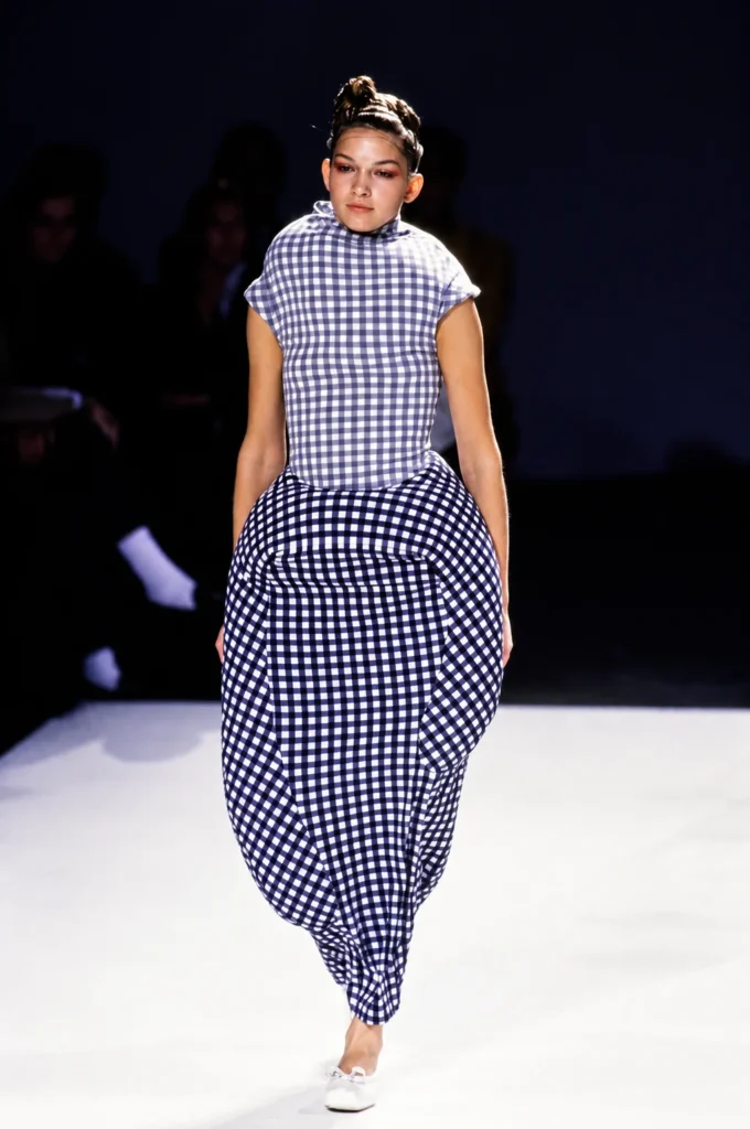 Modelo en pasarela desfilando una pieza de la colección 'Body Meets Dress, Dress Meets Body' (primavera-verano 1997) de Comme des Garçons. El vestido presenta un estampado de cuadros vichy en azul y blanco, destacando por sus formas asimétricas y acolchadas que distorsionan la silueta humana, concepto central de la propuesta vanguardista de Rei Kawakubo.