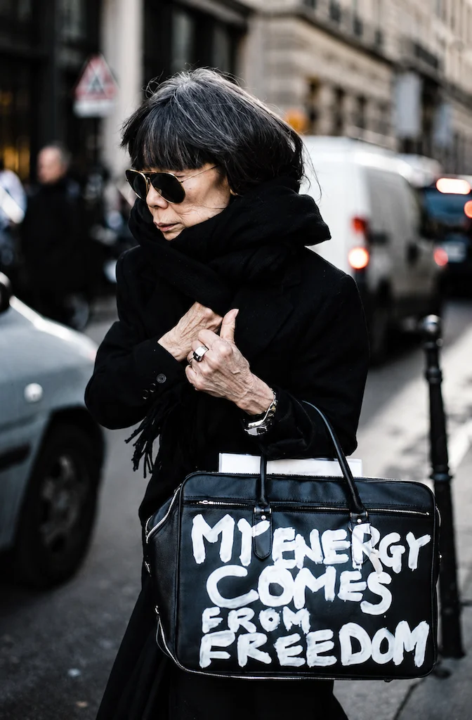 La diseñadora Rei Kawakubo de Comme des Garçons caminando en una vía pública, personificando la moda disruptiva. Viste un abrigo negro minimalista y lleva un maletín de cuero con la leyenda 'My energy comes from freedom'.