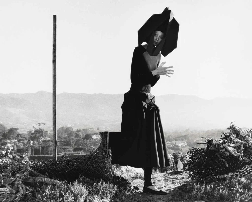 Fotografía editorial en blanco y negro de una modelo en un entorno árido, luciendo un atuendo arquitectónico de Junya Watanabe con un tocado geométrico de gran escala y una falda estructurada de volumen asimétrico.
