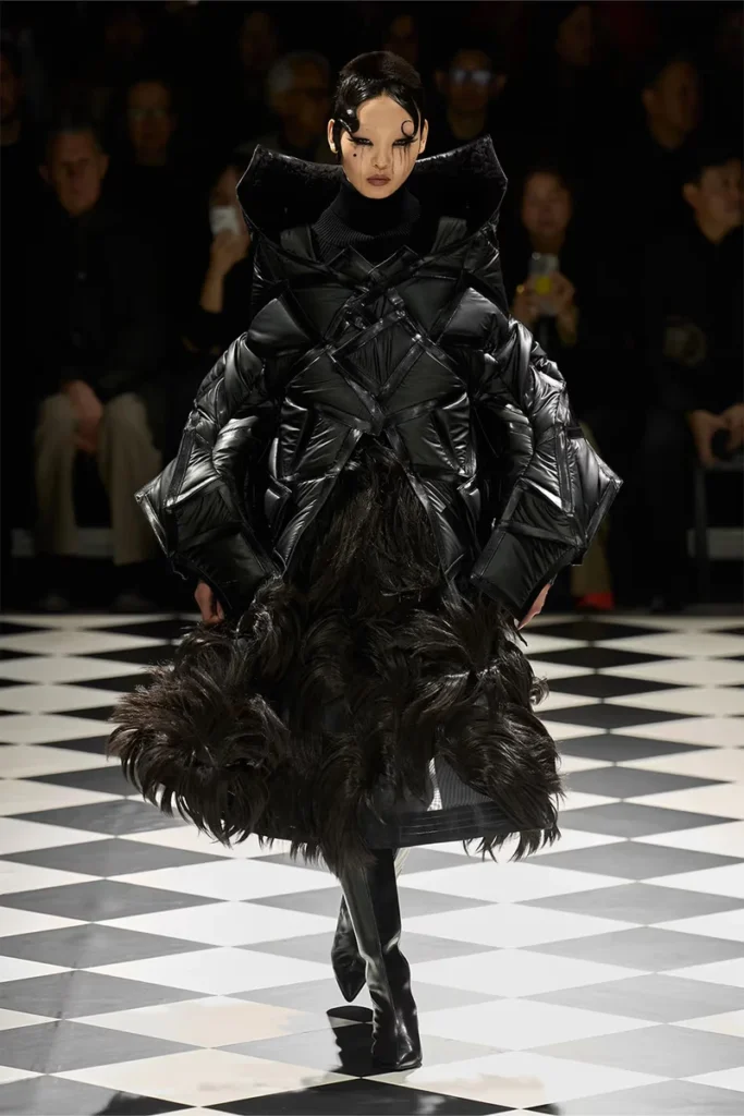 Modelo desfilando sobre un suelo de tablero de ajedrez blanco y negro, luciendo una voluminosa chaqueta acolchada de color negro con un intrincado entramado geométrico en el torso y una falda texturizada con apariencia de pelaje denso.
