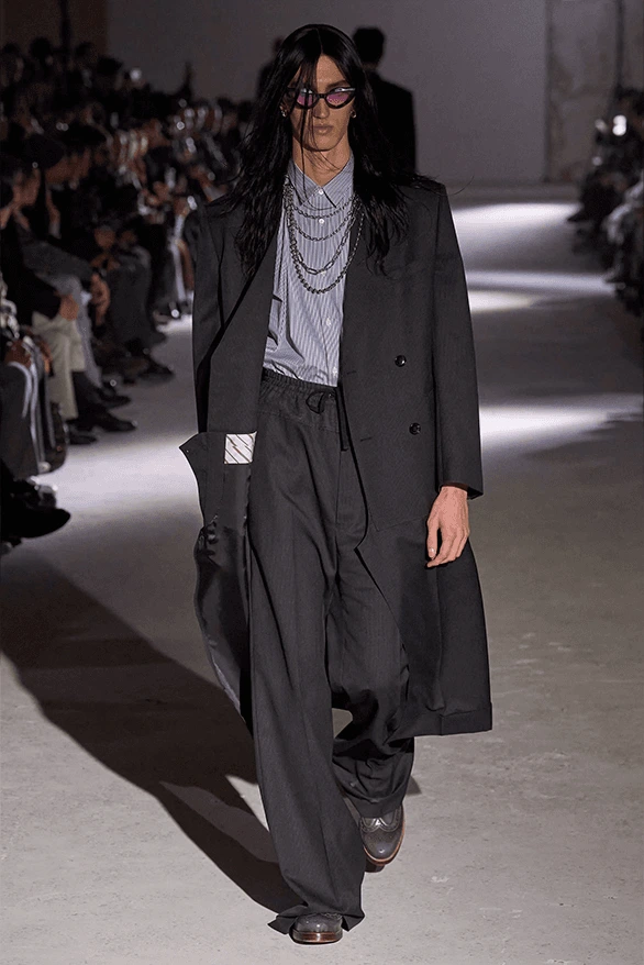 Modelo masculino con cabello largo y gafas oscuras desfilando con un abrigo largo cruzado de sastre en color negro, camisa de rayas finas azules y múltiples capas de cadenas gruesas plateadas al cuello.