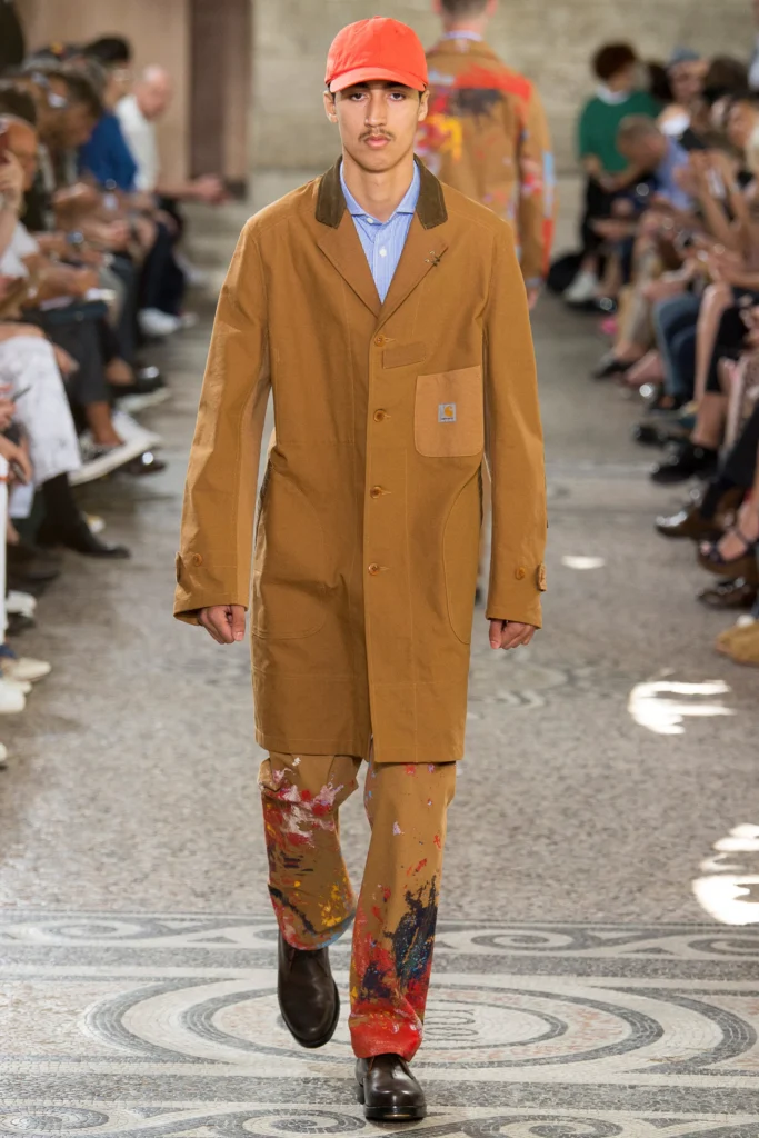 Modelo masculino joven en una pasarela de Junya Watanabe diseñador de modas, vistiendo un abrigo largo de lona color marrón tabaco con cuello de pana y el logotipo de Carhartt en el bolsillo del pecho, combinado con pantalones manchados de pintura multicolor y una gorra naranja brillante.