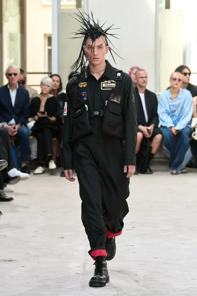 Modelo masculino con un peinado peinado hacia arriba con mechones rígidos y afilados, vistiendo un mono utilitario negro con arneses frontales, hebillas plásticas y parches comerciales, incluyendo logos de Palace y Mastercard de Junya Watanabe diseñador de modas.