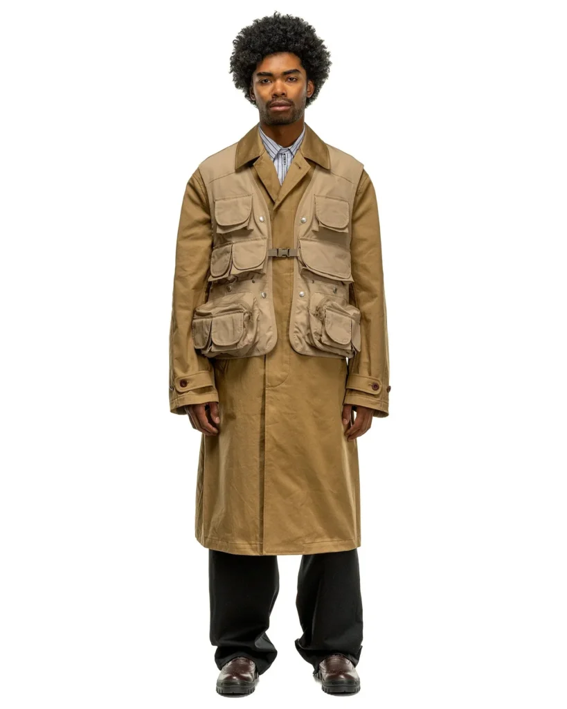 Hombre modelando un trench coat beige de Junya Watanabe MAN con chaleco táctico utilitario integrado de múltiples bolsillos, camisilla de rayas y pantalones negros de corte ancho.