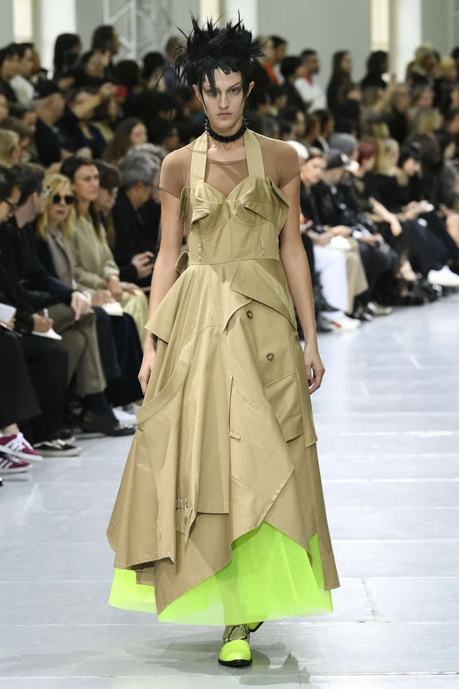Modelo con un peinado punk texturizado y puntiagudo en color negro, vistiendo un vestido asimétrico confeccionado con fragmentos de gabardina beige y un corsé superior, revelando un forro interior de tul verde neón brillante en el bajo deJunya Watanabe diseñador de modas