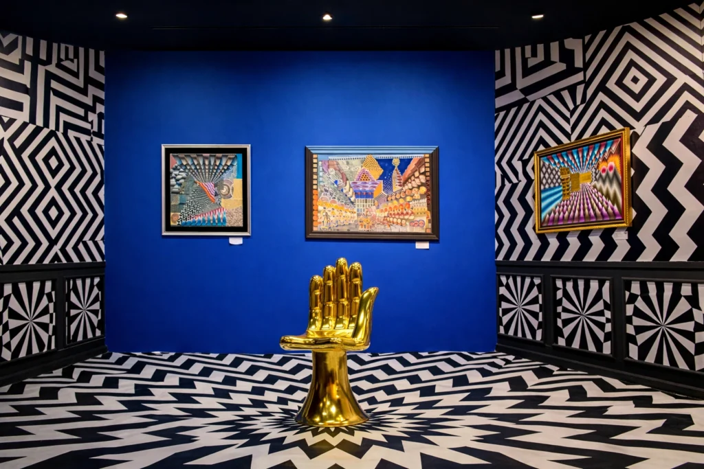 Silla mano dorada centrada frente a un muro azul intenso flanqueado por patrones de laberintos en blanco y negro.