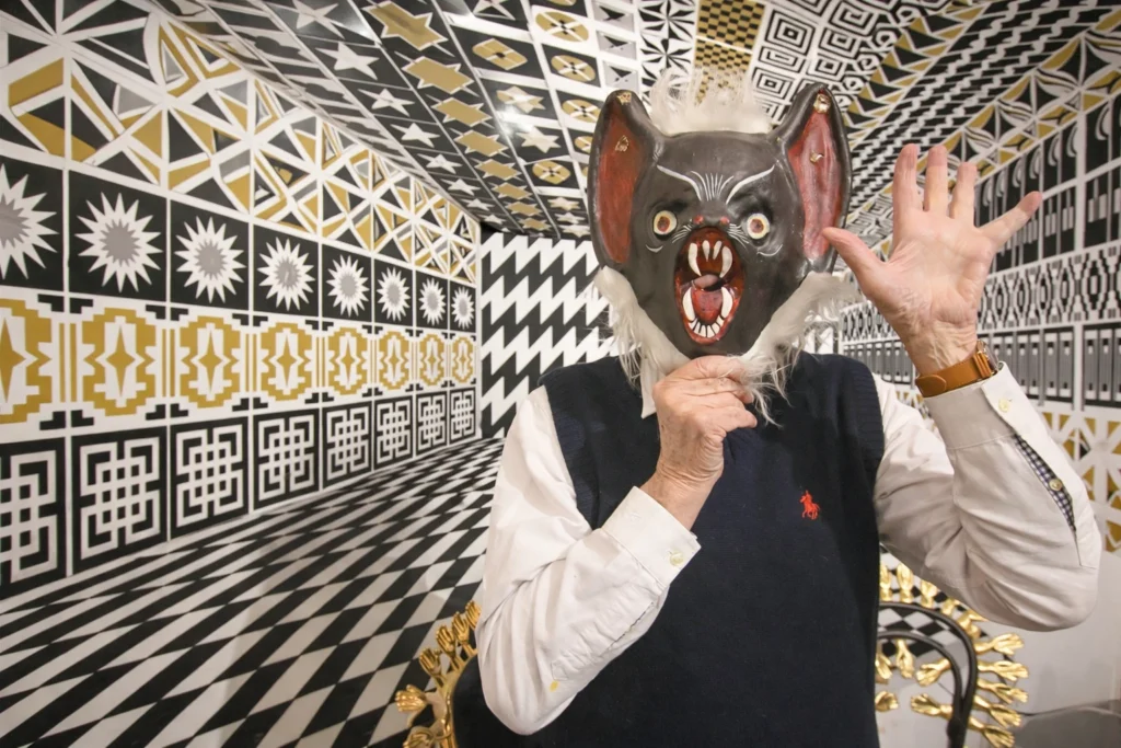 Pedro Friedeberg posando con máscara folclórica frente a un muro de patrones geométricos y op-art.