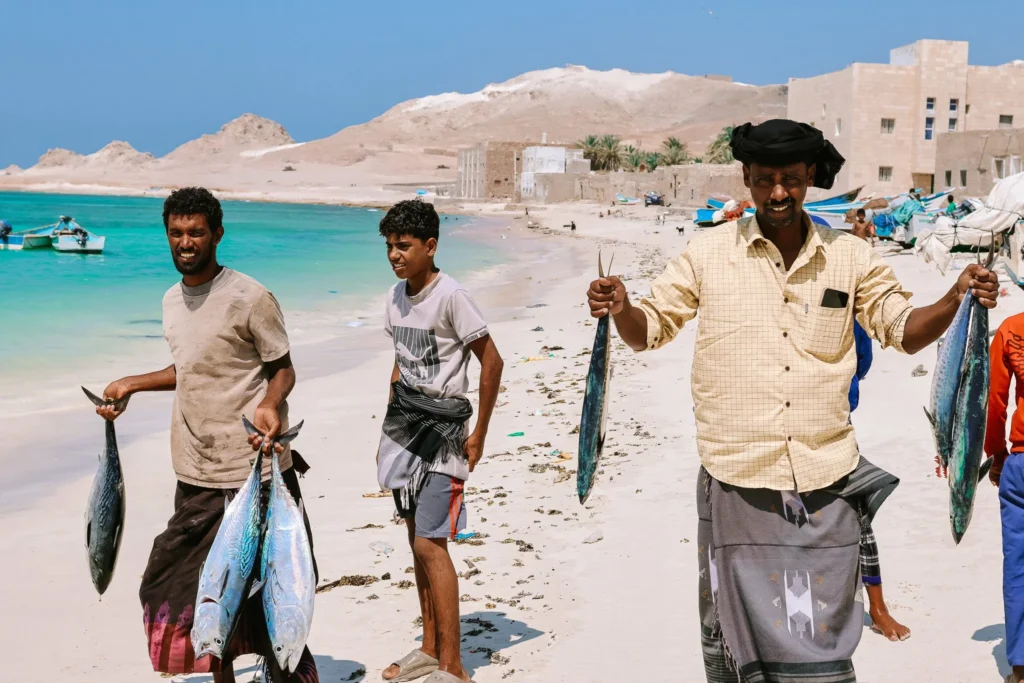 Pescadores locales de la isla de Socotra Yemen sosteniendo pescados atún frescos en una playa de arena blanca con botes al fondo.