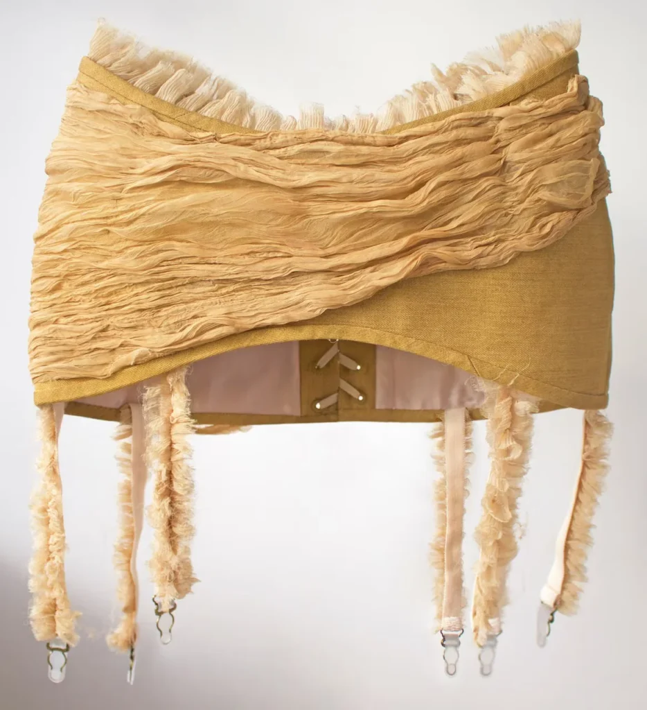 Corsé en tonos ocre y dorado de Michaela Stark con detalles de volantes y ligueros, resaltando la textura de la tela y el diseño de alta costura conceptual.