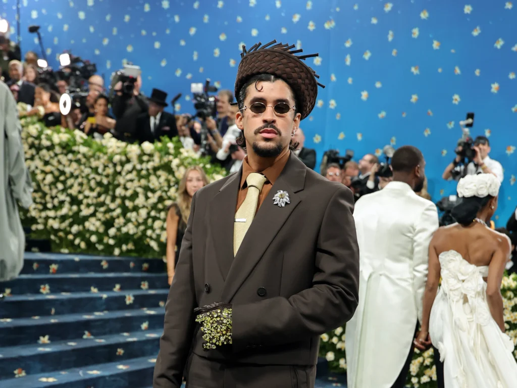 Bad Bunny con traje marrón, gafas de sol y sombrero de estructura tejida posando frente a un fondo azul con flores.