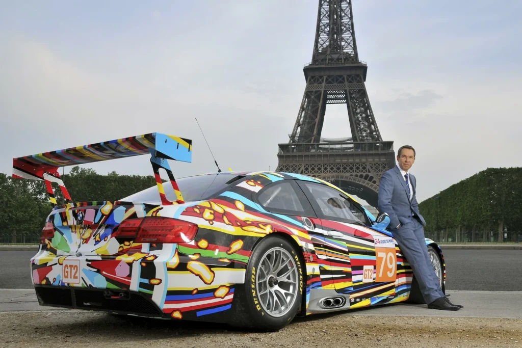 El artista Jeff Koons posando junto al BMW M3 GT2 Art Car en París, con la Torre Eiffel de fondo, destacando el diseño de líneas de colores explosivas que simulan velocidad y energía.
