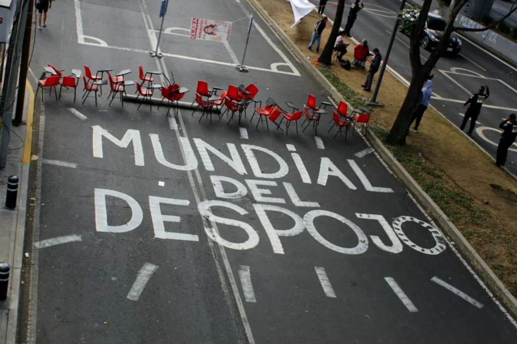 Pinta sobre el asfalto de una avenida con la frase "Mundial del Despojo" en letras blancas grandes, bloqueada por una fila de pupitres escolares rojos en señal de protesta.