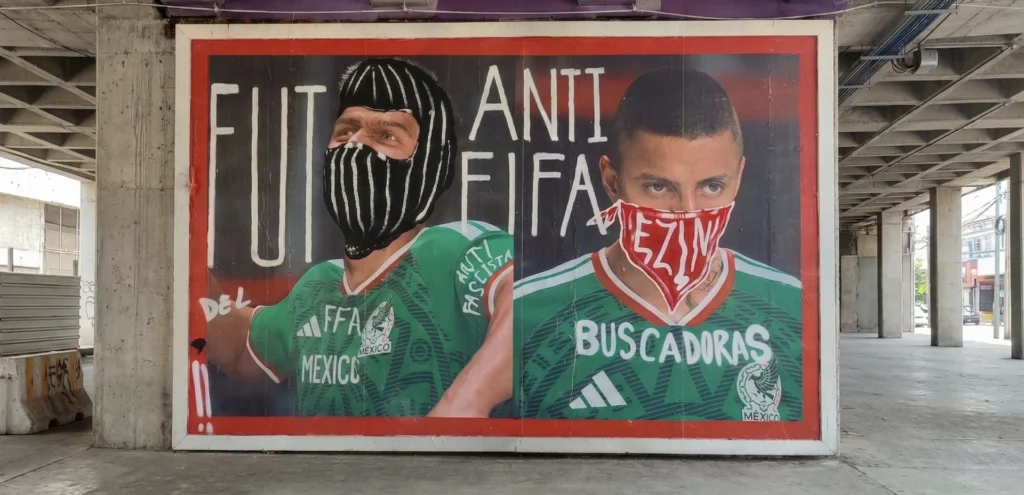 Mural urbano con la leyenda "Anti FIFA" y "Buscadoras", mostrando a dos futbolistas con el rostro cubierto por pasamontañas y paliacates en un entorno de protesta social.