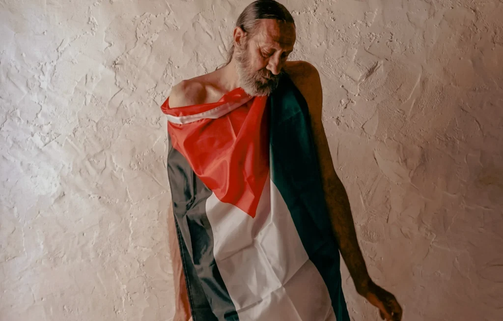 Retrato de alta resolución de Miguel Adrover en su refugio de Mallorca, envuelto en una bandera de Palestina, simbolizando su postura biopolítica y su negativa a separar el arte del compromiso humanitario.