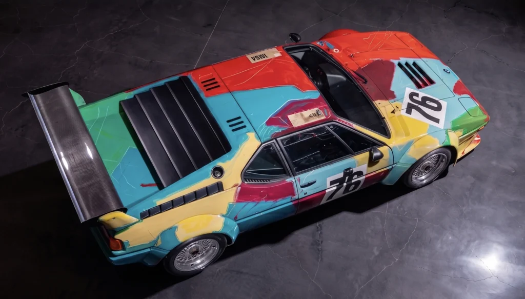 Vista aérea cenital del BMW M1 Art Car pintado por Andy Warhol, mostrando la distribución de colores vibrantes, pinceladas gestuales y el número 76 sobre el capó y puertas en un entorno de estudio.