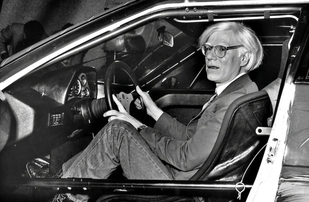 Andy Warhol sentado en el asiento del conductor del BMW M1 Art Car en 1979, capturando el momento en que el artista se integra con su obra de ingeniería.