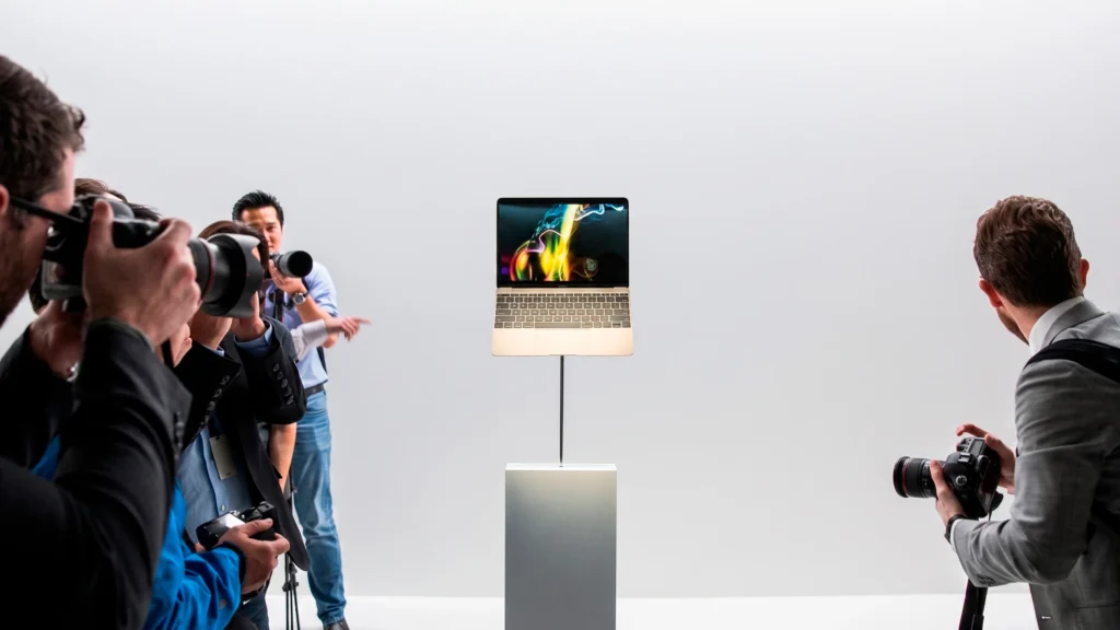 Fotografía de prensa que muestra a múltiples fotógrafos rodeando y fotografiando una MacBook dorada sobre un pedestal, ilustrando la mística y el fetiche de hardware que Apple debe recuperar frente a la competencia.
