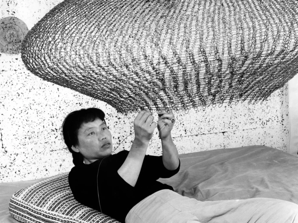 Fotografía en blanco y negro de Ruth Asawa trabajando en una escultura de alambre suspendida mientras descansa, ilustrando su método de tejido manual y paciencia meditativa.