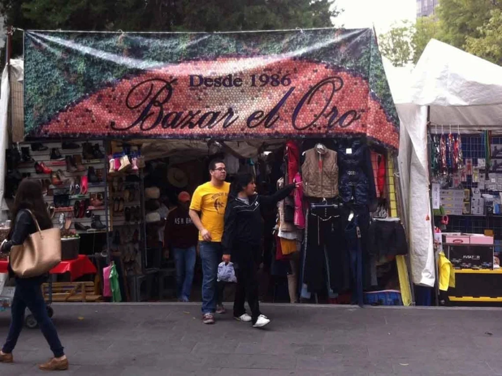 Fachada de un puesto ambulante con lona publicitaria que dice Bazar el Oro Desde 1986, mostrando ropa y calzado en la Colonia Roma Norte de la CDMX.
