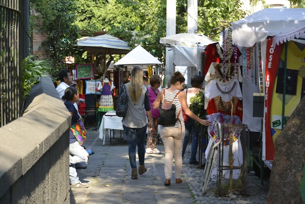 Visitantes caminando junto a puestos de ropa típica bordada y artesanías en el andador empedrado del Bazar Sábado en San Ángel, CDMX.