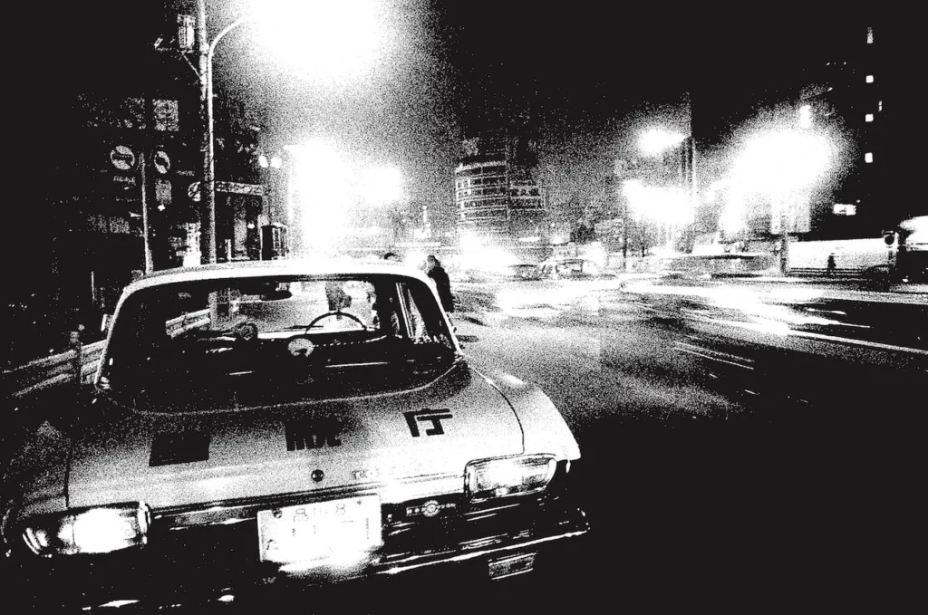 Fotografía urbana en blanco y negro de Daidō Moriyama que muestra la parte trasera de un automóvil clásico de la Policía Metropolitana de Tokio con letras en japonés en la cajuela, circulando de noche en estilo are bure bokeh por una avenida iluminada en 1969.