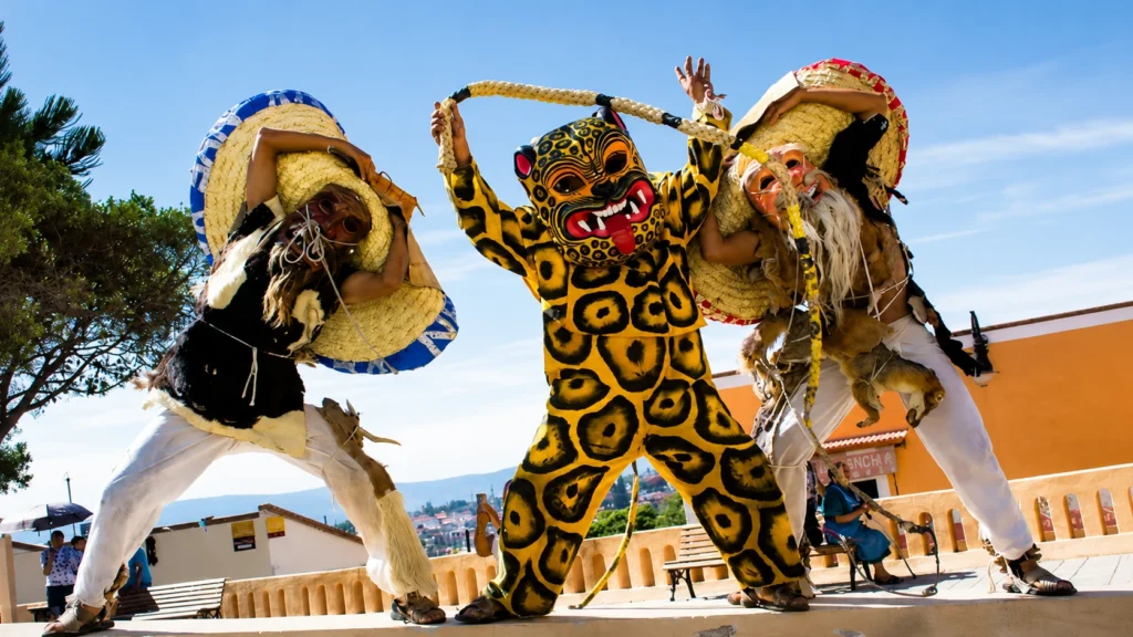 Tres danzantes de la Danza de los Tecuanes con trajes de jaguar y viejos cazadores posando con máscaras y sombreros gigantes de paja bajo un cielo azul.