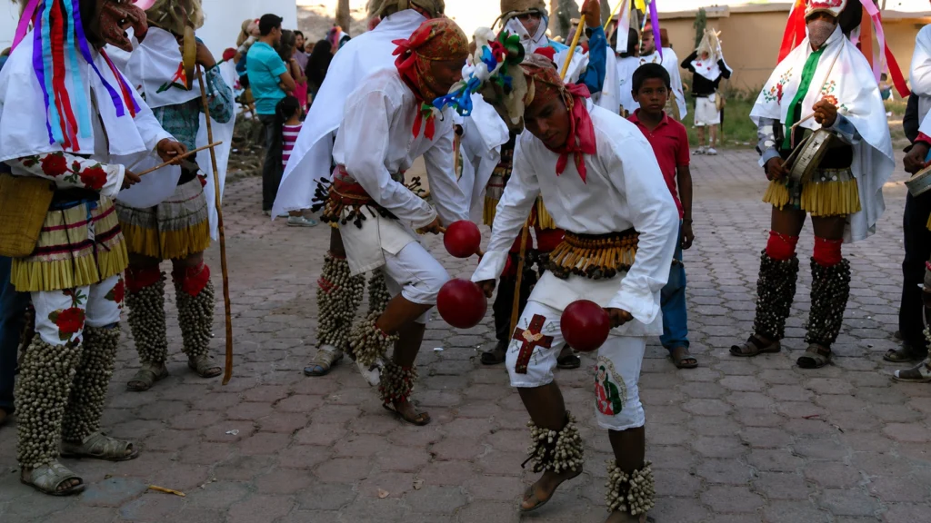 Danzantes tradicionales de la Danza del Venado inclinados tocando sonajas rojas con tenábaris en las piernas durante una celebración comunitaria.