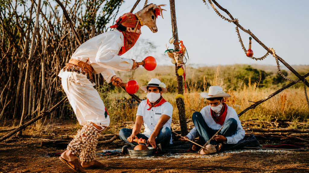 Danzante con cabeza de venado tocando sonajas rojas junto a músicos tradicionales sentados que tocan el tambor de agua en un entorno rural.