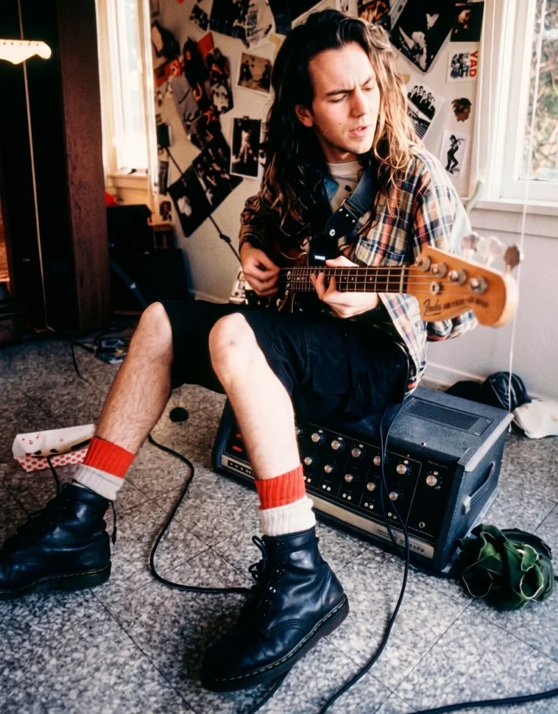 Eddie Vedder de Pearl Jam tocando el bajo sobre un amplificador en un departamento de Seattle, vistiendo camisa de franela, shorts y botas Dr. Martens, ejemplificando el diseño de producción y vestuario de la película Singles.