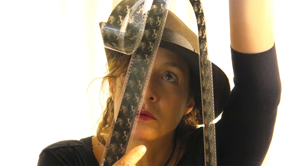 La cineasta mexicana Ximena Cuevas sosteniendo y revisando a contraluz una tira de película cinematográfica de celuloide frente a sus ojos.