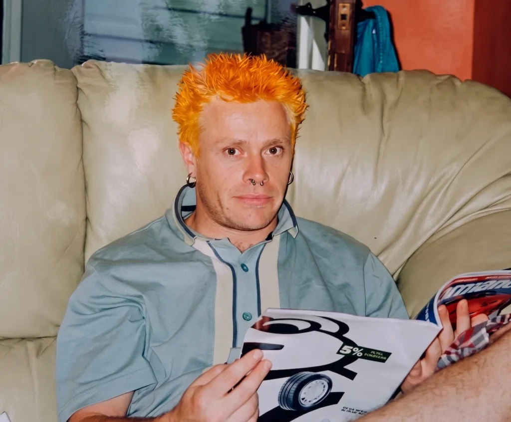 Keith Flint sentado en un sofá sosteniendo una revista, representando su faceta humana y su arraigo a la cultura de clase trabajadora fuera de los escenarios.