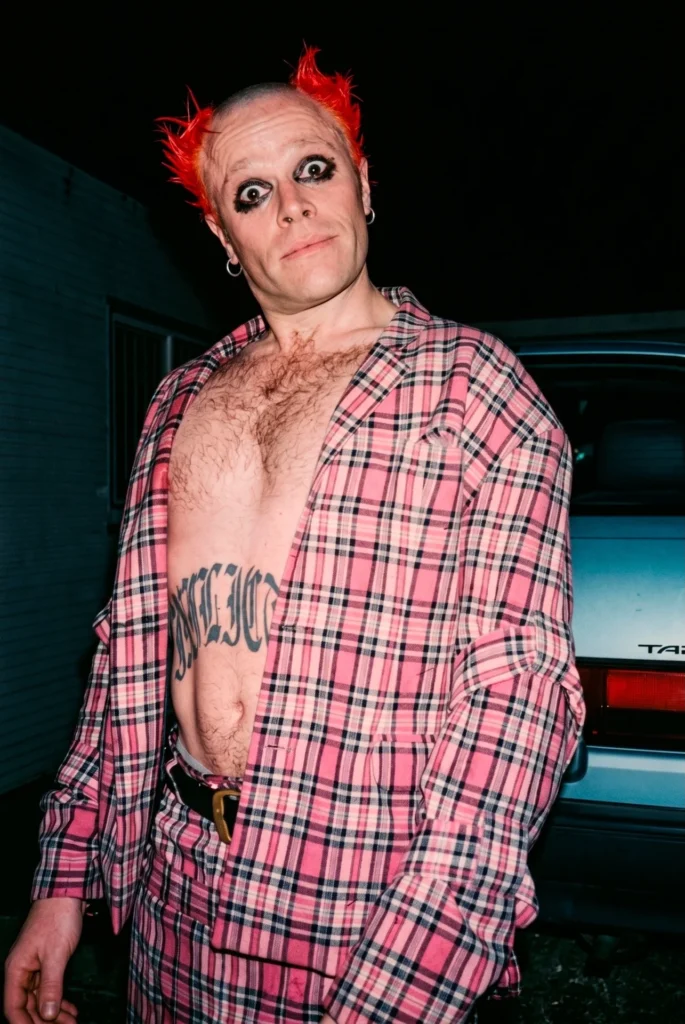 Keith Flint con cabello rojo tipo 'devil horns', saco de cuadros rosa y tatuaje abdominal, capturando el sabotaje estético al pop de los 90.