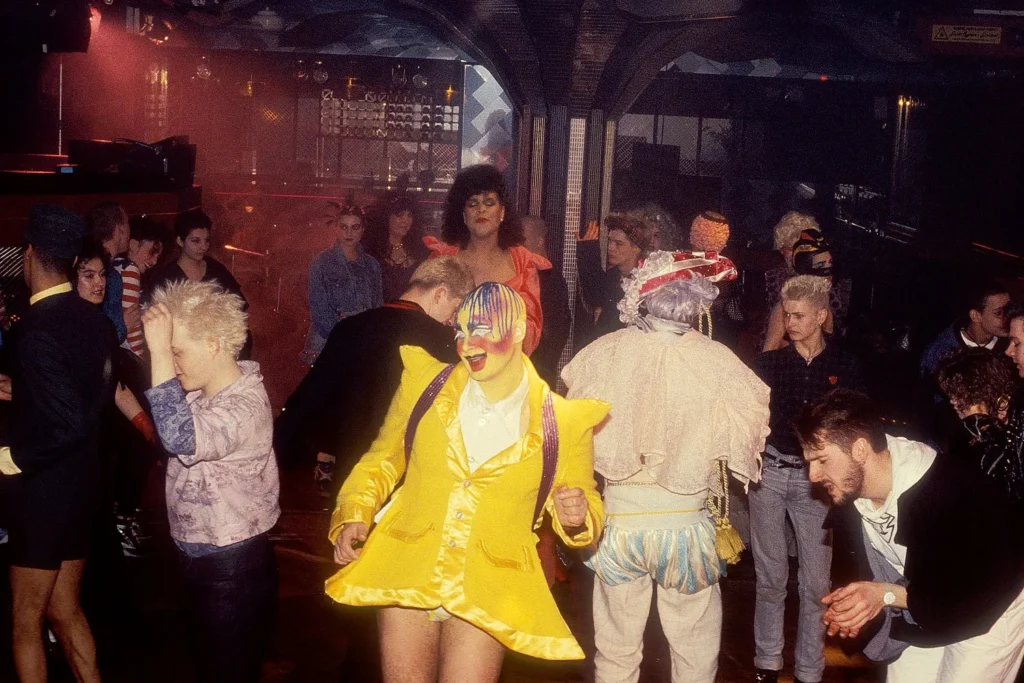 El artista de performance Leigh Bowery baila en la pista del club nocturno Taboo vistiendo un saco amarillo satinado y maquillaje facial de colores, rodeado de personas en una fiesta clandestina de Londres en 1985.