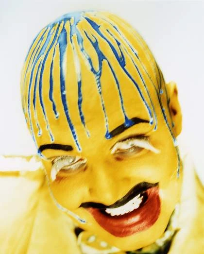 Primer plano de estudio del artista Leigh Bowery con el rostro pintado de amarillo brillante, pegamento azul goteando sobre su cabeza calva, cejas negras marcadas, pestañas de hilos blancos y labios pintados, fotografiado por Nick Knight en 1992.