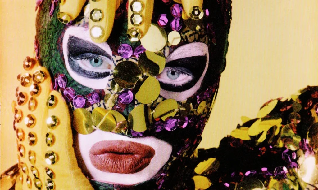 Primer plano del artista Leigh Bowery usando una máscara completa cubierta de lentejuelas moradas, doradas y verdes, guantes amarillos con incrustaciones de pedrería y un maquillaje de labios delineado de forma exagerada.