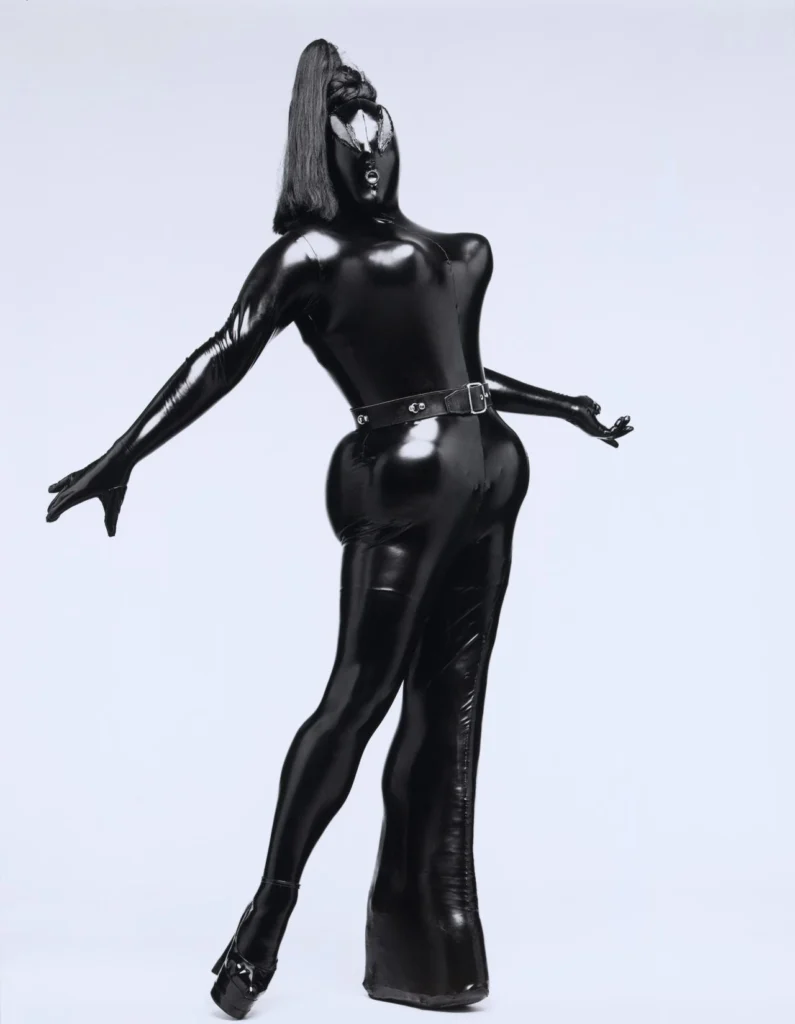 El artista de performance Leigh Bowery posa de cuerpo entero usando un traje de látex negro brillante ajustado con corsé extremo, cinturón, máscara facial total y una coleta de cabello negro, fotografiado por Fergus Greer en 1988.