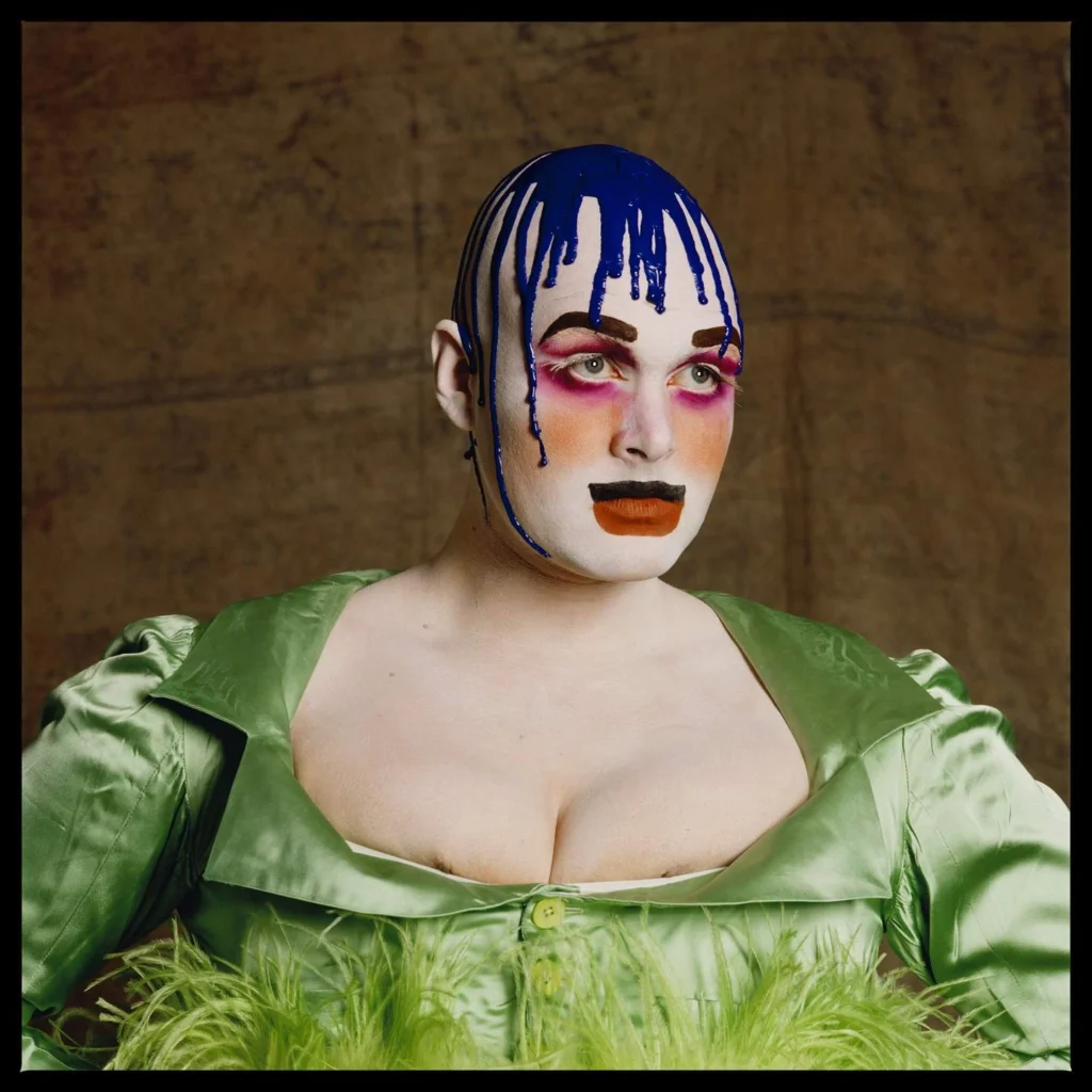 El artista de performance Leigh Bowery posa de frente con un vestido verde satinado de escote profundo con plumas, el rostro pintado de blanco con maquillaje dramático y pegamento azul derretido goteando por su cabeza calva, retratado por Fergus Greer.