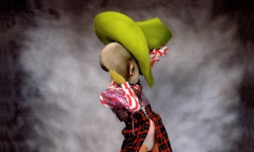 El artista de performance Leigh Bowery posa de perfil usando un enorme sombrero verde de ala ancha, una máscara gris ajustada que deforma su cabeza, y un traje de falda abierta a cuadros rojos con corsé, fotografiado por Fergus Greer.